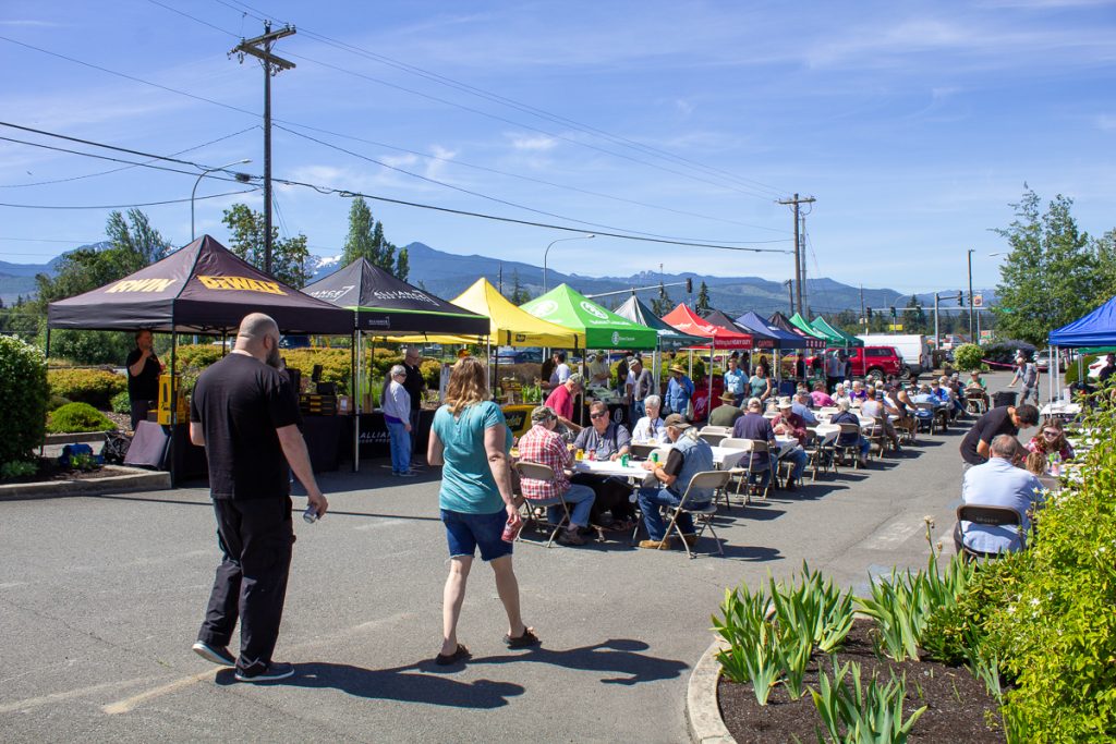 People enjoying food at tables surrounded by vendor booths with a couple walking down the center on a clear day with mountains in the background.